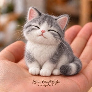 Può includere: Una piccola statuetta di gatto in lana infeltrita con pelo grigio e bianco, orecchie rosa e un'espressione sorridente. Il gatto ha lunghi baffi bianchi ed è seduto in una posizione rilassata. Il testo LunaCraft Gifts è stampato nell'immagine.