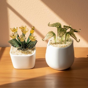 May include: Two small, white ceramic pots with glass plant sculptures. One pot features yellow flower sculptures with green leaves, and the other has green leaf sculptures. Both pots are filled with small, light-colored pebbles.