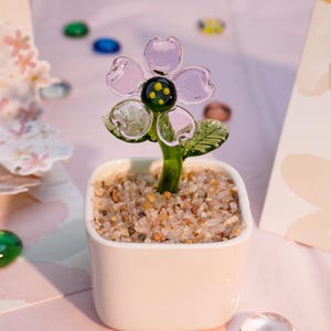 May include: A glass flower sculpture in a white square pot filled with small pebbles. The flower has translucent pink petals, a black centre with yellow dots, and green leaves and stem. The sculpture is on a pink surface.