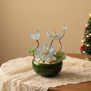 May include: A decorative floral arrangement featuring blue glass lotus flowers with yellow centres and green leaves. The flowers are in a dark green ceramic bowl filled with white stones. The arrangement sits on a lace tablecloth.