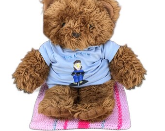 Op de afbeelding: Een bruine teddybeer met een lichtblauw t-shirt met de tekst "Venus Aqueous" en een afbeelding van een persoon. De beer zit op een roze en wit gestreepte gebreide deken.