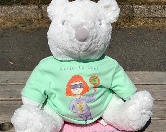 Op de afbeelding: Een witte teddybeer draagt een lichtgroen t-shirt met de tekst "Reflecto Girl" en een cartoonillustratie van een superheldin die een lolly vasthoudt. De beer zit op een roze en wit gestreepte gebreide deken.