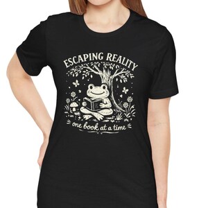 May include: Black t-shirt with a white graphic of a frog reading a book under a tree. The text "ESCAPING REALITY" is above the frog, and "one book at a time" is below.