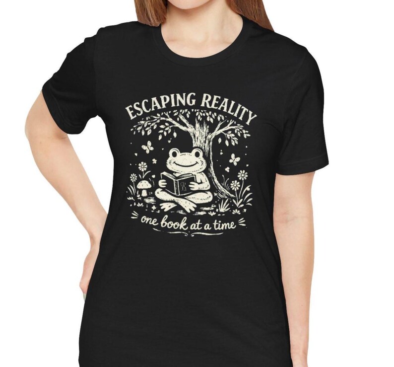 May include: Black t-shirt with a white graphic of a frog reading a book under a tree. The text "ESCAPING REALITY" is above the frog, and "one book at a time" is below.