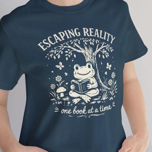 May include: Navy blue t-shirt with a whimsical graphic. The design features a smiling frog reading a book under a tree, with the words "ESCAPING REALITY" and "one book at a time" in white script. Butterflies and flowers surround the frog.