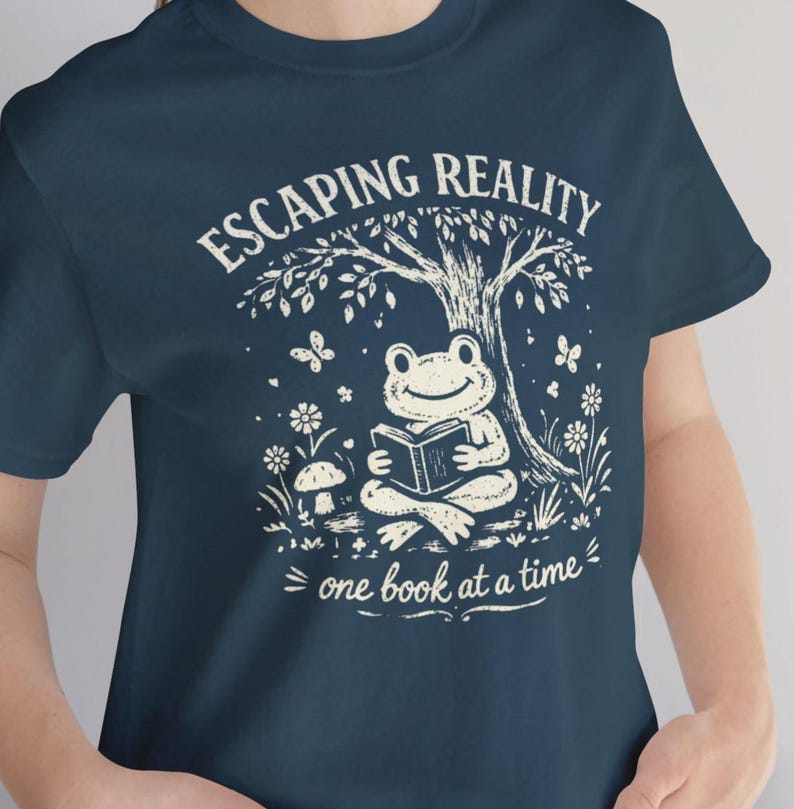 May include: Navy blue t-shirt with a whimsical graphic. The design features a smiling frog reading a book under a tree, with the words "ESCAPING REALITY" and "one book at a time" in white script. Butterflies and flowers surround the frog.