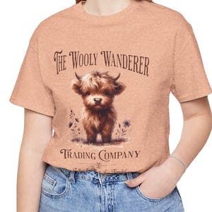 May include: Peach-colored t-shirt featuring a watercolor illustration of a Highland cow calf. The text "The Wooly Wanderer Trading Company" is printed above and below the image. The shirt is made of a soft material.