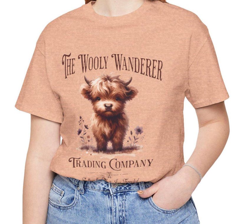 May include: Peach-colored t-shirt featuring a watercolor illustration of a Highland cow calf. The text "The Wooly Wanderer Trading Company" is printed above and below the image. The shirt is made of a soft material.