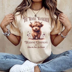 May include: Cream-colored t-shirt featuring the text "The Wooly Wanderer Trading Company" and a brown Highland calf illustration. The shirt also includes the phrase "Follow the Trails Less Traveled" in a vintage style.