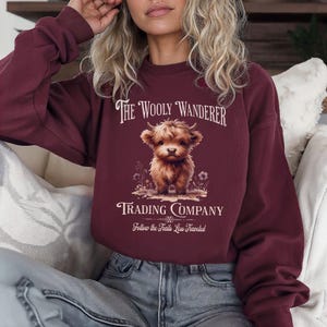 May include: Burgundy-colored sweatshirt featuring the text "The Wooly Wanderer Trading Company" and a graphic of a Highland cow. The design includes the phrase "Follow the Trails Less Traveled" below the cow illustration.