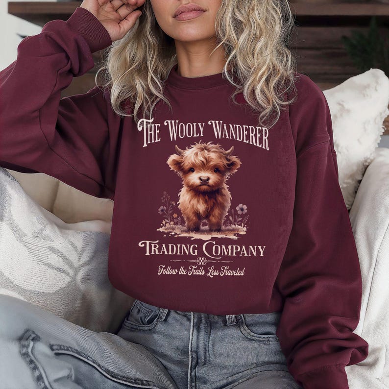 May include: Burgundy-colored sweatshirt featuring the text "The Wooly Wanderer Trading Company" and a graphic of a Highland cow. The design includes the phrase "Follow the Trails Less Traveled" below the cow illustration.
