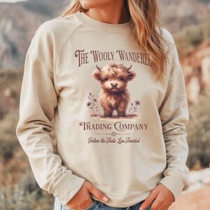 May include: Beige long-sleeve sweatshirt with a graphic of a Highland cow and the text "The Wooly Wanderer Trading Company". The design includes purple flowers and the phrase "Follow the Trails Less Traveled".