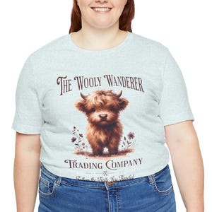 May include: Light gray t-shirt featuring a brown Highland cow illustration with the text "The Wooly Wanderer Trading Company." The design includes floral accents and the phrase "Follow the Trails Less Traveled."