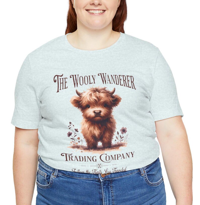 May include: Light gray t-shirt featuring a brown Highland cow illustration with the text "The Wooly Wanderer Trading Company." The design includes floral accents and the phrase "Follow the Trails Less Traveled."