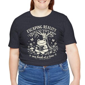 May include: Dark gray t-shirt with the text "ESCAPING REALITY" above an illustration of a frog reading a book under a tree. The text "one book at a time" is below the frog.