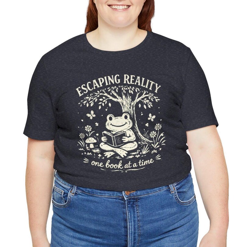 May include: Dark gray t-shirt with the text "ESCAPING REALITY" above an illustration of a frog reading a book under a tree. The text "one book at a time" is below the frog.