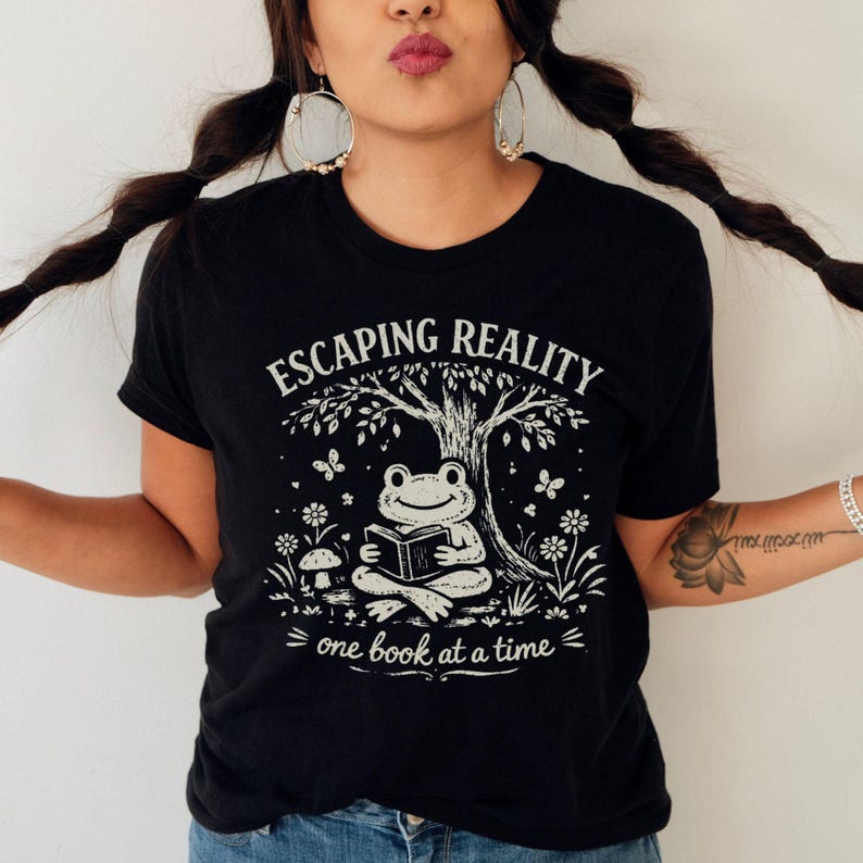 May include: Black t-shirt with a white graphic of a frog reading under a tree, with the text "ESCAPING REALITY" above and "one book at a time" below. The design includes butterflies and flowers.