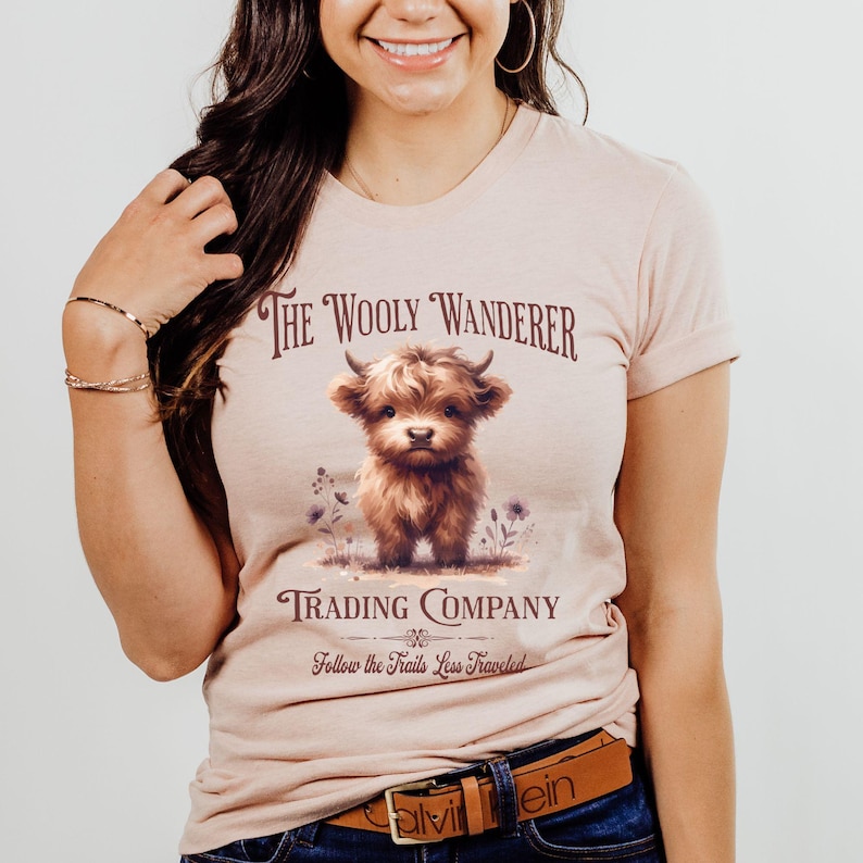 May include: A light pink t-shirt featuring a watercolor illustration of a Highland cow calf. The text above the calf reads "The Wooly Wanderer Trading Company" and below it reads "Follow the Trails Less Traveled".