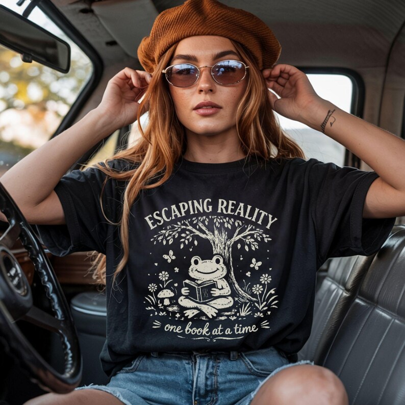May include: Black t-shirt with the text "Escaping Reality" and a graphic of a frog reading under a tree. The design includes butterflies, flowers, and the phrase "one book at a time". The person in the image is wearing a brown beret and sunglasses.