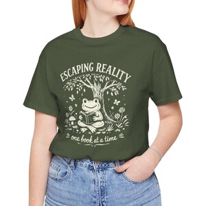 May include: Olive green t-shirt with a whimsical graphic. The design features a frog reading a book under a tree, with the text "Escaping Reality, one book at a time" in white. The shirt is a casual, comfortable style.