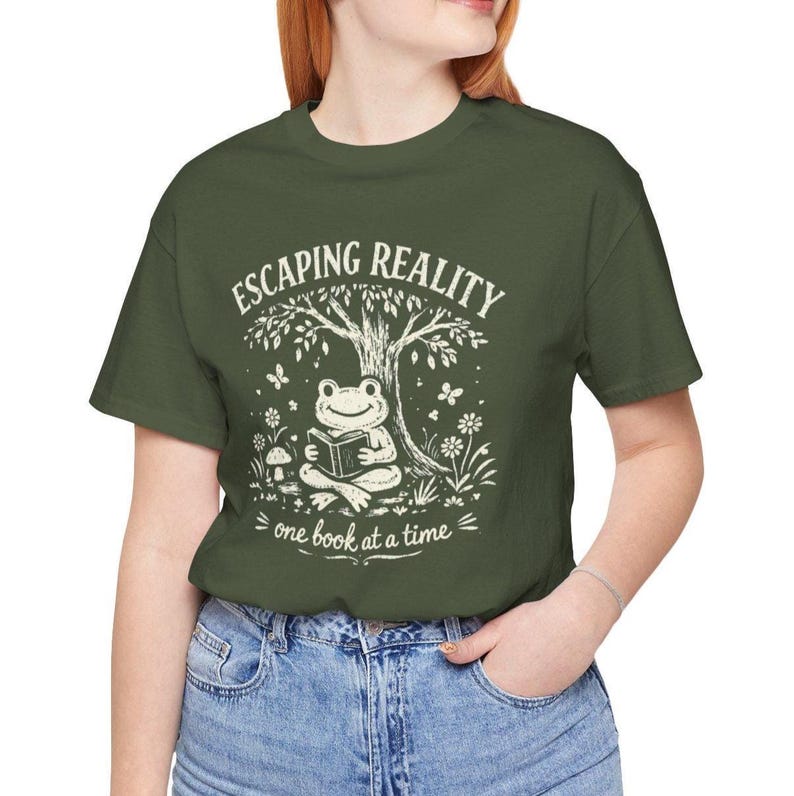 May include: Olive green t-shirt with a whimsical graphic. The design features a frog reading a book under a tree, with the text "Escaping Reality, one book at a time" in white. The shirt is a casual, comfortable style.