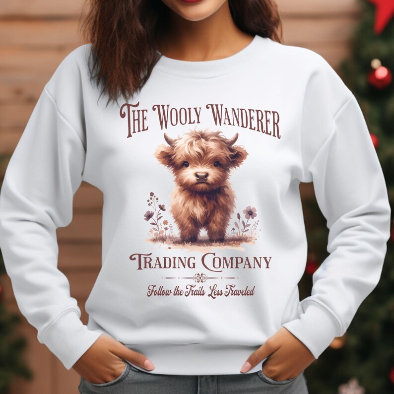 May include: White sweatshirt featuring a brown illustration of a Highland cow with small horns. The text "The Wooly Wanderer Trading Company" and "Follow the Trails Less Traveled" are printed above and below the cow graphic.