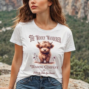 May include: White t-shirt featuring a brown Highland cow illustration with the text "The Wooly Wanderer Trading Company". The shirt also includes the phrase "Follow the Trails Less Traveled" in a decorative font.