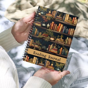 May include: A spiral bound journal with a dark blue cover featuring a vintage illustration of a bookshelf filled with books. The cover has a gold banner with the text "Reading Journal".