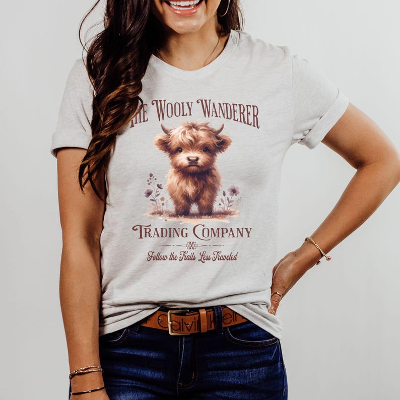 May include: A light gray t-shirt featuring a brown Highland calf illustration with the text "The Wooly Wanderer Trading Company". The shirt also includes the phrase "Follow the Trails Less Traveled".