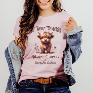 May include: A light pink t-shirt featuring a brown Highland cow illustration with the text "The Wooly Wanderer Trading Company" and "Follow the Trails Less Traveled". The shirt has a crew neck and short sleeves.