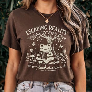 May include: Brown t-shirt with a whimsical graphic. The design features a frog reading a book under a tree, with the text "ESCAPING REALITY one book at a time." Butterflies and flowers surround the frog. The shirt is a casual, comfortable style.