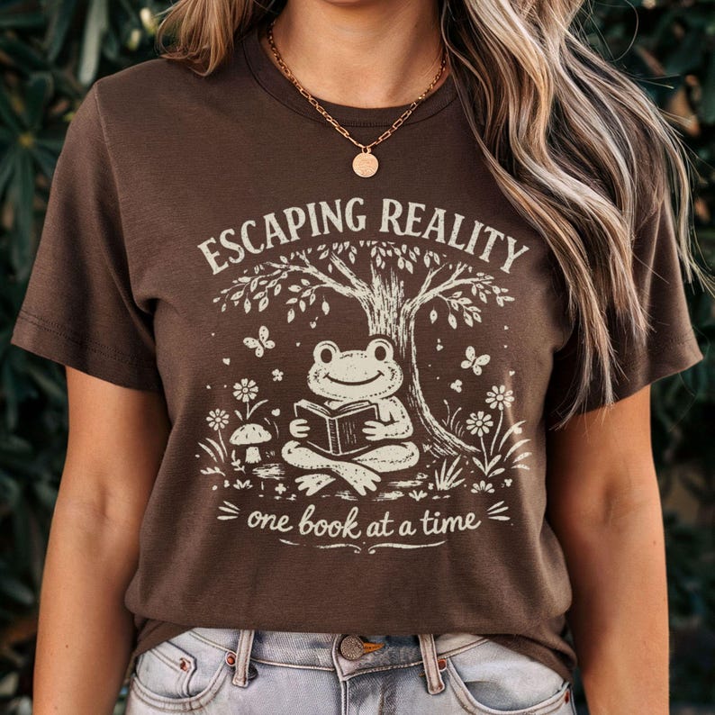May include: Brown t-shirt with a whimsical graphic. The design features a frog reading a book under a tree, with the text "ESCAPING REALITY one book at a time." Butterflies and flowers surround the frog. The shirt is a casual, comfortable style.