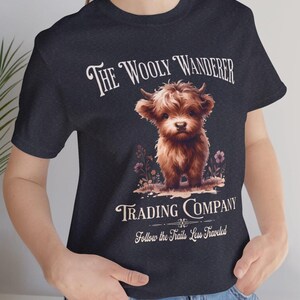 May include: Dark navy blue t-shirt featuring a brown Highland calf illustration. The text reads "The Wooly Wanderer Trading Company" with the tagline "Follow the Trails Less Traveled". The calf is standing in a field of flowers.