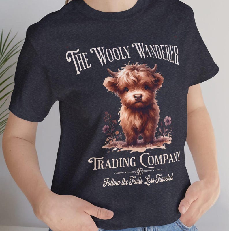 May include: Dark navy blue t-shirt featuring a brown Highland calf illustration. The text reads "The Wooly Wanderer Trading Company" with the tagline "Follow the Trails Less Traveled". The calf is standing in a field of flowers.