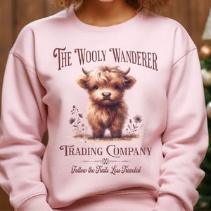 May include: A light pink sweatshirt featuring the text "The Wooly Wanderer Trading Company" with a graphic of a fluffy brown Highland cow. The phrase "Follow the Trails Less Traveled" is printed below the graphic.