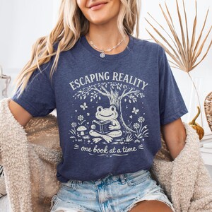 May include: A navy blue t-shirt with a whimsical graphic. The design features a frog reading a book under a tree, with the words "Escaping Reality" and "one book at a time". The shirt is a comfortable, casual style.