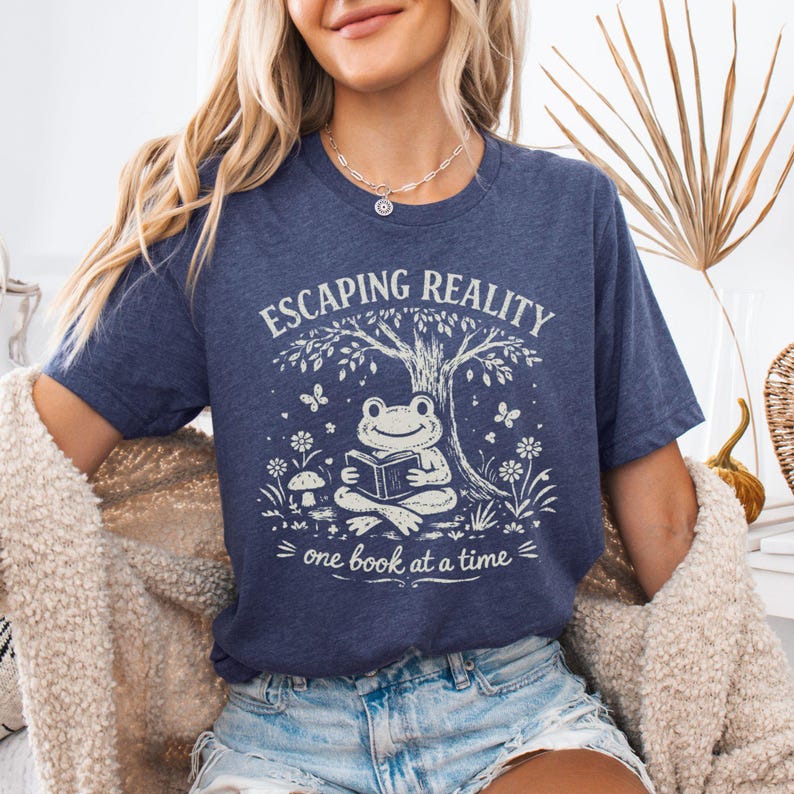 May include: A navy blue t-shirt with a whimsical graphic. The design features a frog reading a book under a tree, with the words "Escaping Reality" and "one book at a time". The shirt is a comfortable, casual style.