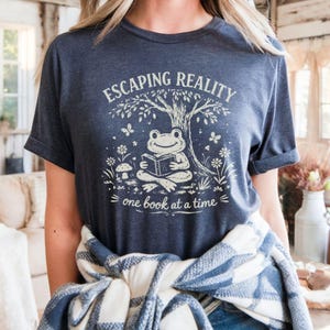 May include: Dark blue t-shirt with the text "ESCAPING REALITY" above an illustration of a frog reading a book under a tree. The phrase "one book at a time" is below the illustration. The shirt has rolled-up sleeves.