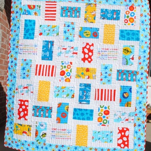 May include: A white quilt with a blue and yellow border. The quilt is made up of various squares with different patterns, including red and white stripes, yellow polka dots, and blue and white fish. The text "THE CAT IN THE HAT" is visible on one of the squares.