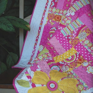 Peut inclure: Une courtepointe faite à la main avec une grande fleur jaune appliquée et des accents roses et verts. La courtepointe présente un motif patchwork avec divers motifs roses, y compris des pois et des imprimés floraux. La bordure est blanche avec une bordure rose.