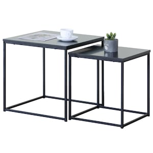 May include: Two nesting side tables with dark grey tops and black metal frames. A white teacup and saucer sit on the larger table, and a small potted plant is on the smaller table. The tables are square and have a modern design.