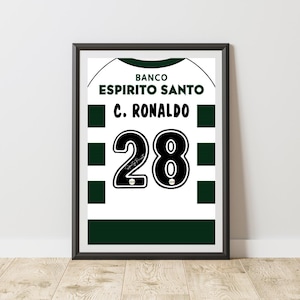 May include: Framed print of a soccer jersey. The white jersey features green accents, the text "BANCO ESPIRITO SANTO", and "C. RONALDO" with the number "28" in bold black. The frame is black.
