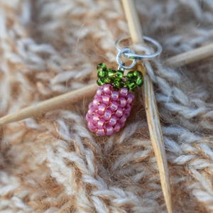 Pode incluir: Um pequeno pendente tridimensional em forma de framboesa, feito de pequenas contas de vidro cor-de-rosa e verdes. O pendente está preso a um anel de metal prateado, posicionado à volta de uma agulha de tricot de madeira clara.
