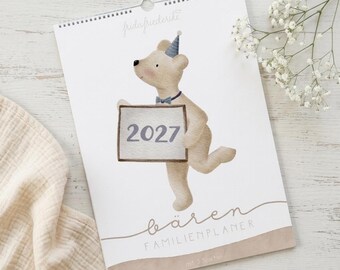 Peut inclure: Un calendrier mural avec une illustration à l'aquarelle d'un ours brun clair portant un chapeau de fête bleu et un nœud papillon, tenant un panneau indiquant 2027. Le texte sous l'ours indique bären FAMILIENPLANER et fridafriederike.
