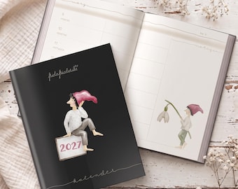 May include: A black 2027 calendar with a graphic of a figure in a pink hat sitting on a sign, placed over an open planner showing weekly pages with German text and a small illustration of a figure holding a white flower.