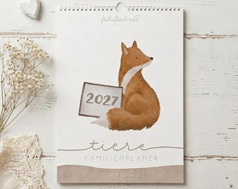 Peut inclure: Un calendrier mural pour 2027 intitulé tiere FAMILIENPLANER avec 5 Spalten. La couverture présente une illustration à l'aquarelle d'un renard brun et blanc tenant un panneau indiquant 2027. La marque fridafriederike est imprimée en haut.