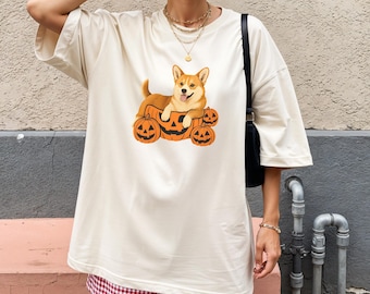 May include: Cream-colored t-shirt featuring a cartoon corgi dog resting on a pumpkin carriage with three jack-o'-lanterns. The dog is light brown and white. The pumpkins are orange. The shirt is paired with red and white checkered shorts.