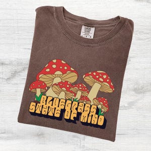 May include: Brown t-shirt with a graphic of red and white mushrooms and the text "Bluegrass State of Mind" in a retro font. The shirt is folded and has a Comfort Colors tag.