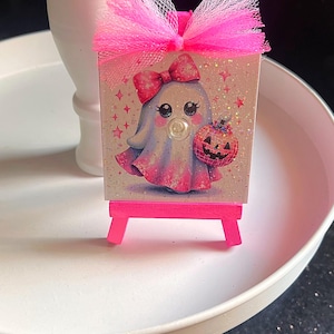 May include: A pink mini easel holds a small canvas featuring a cartoon ghost with a bow, holding a jack-o'-lantern. The canvas is topped with a pink tulle bow. The easel and canvas have glitter accents, creating a festive Halloween decoration.