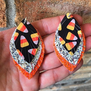 May include: A pair of dangle earrings with a layered design. The top layer features a black fabric with a white and orange candy corn pattern. The middle layer is silver glitter and the bottom layer is orange glitter.
